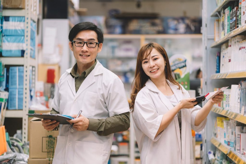 Two pharmacists: one is holding clip board and smiling, one is holding medication from shelf and smiling